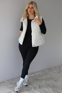 Woman wearing a light sand colored puffer vest with a fitted black long sleeved top and fitted leggings with high white ankle socks and silver sneakers. The photo is taken indoors against a white wall with white marble flooring. 