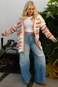 Woman model wearing an orange, lavender, yellow, muted green, blue, and white striped oversized cardigan with functional button closures in the front and an oversized fit with a light wash flared hem denim. The photo is taken outdoors against a white wall and greenery in the back. The photo shows the cardigan unbuttoned with a white cropped top underneath.