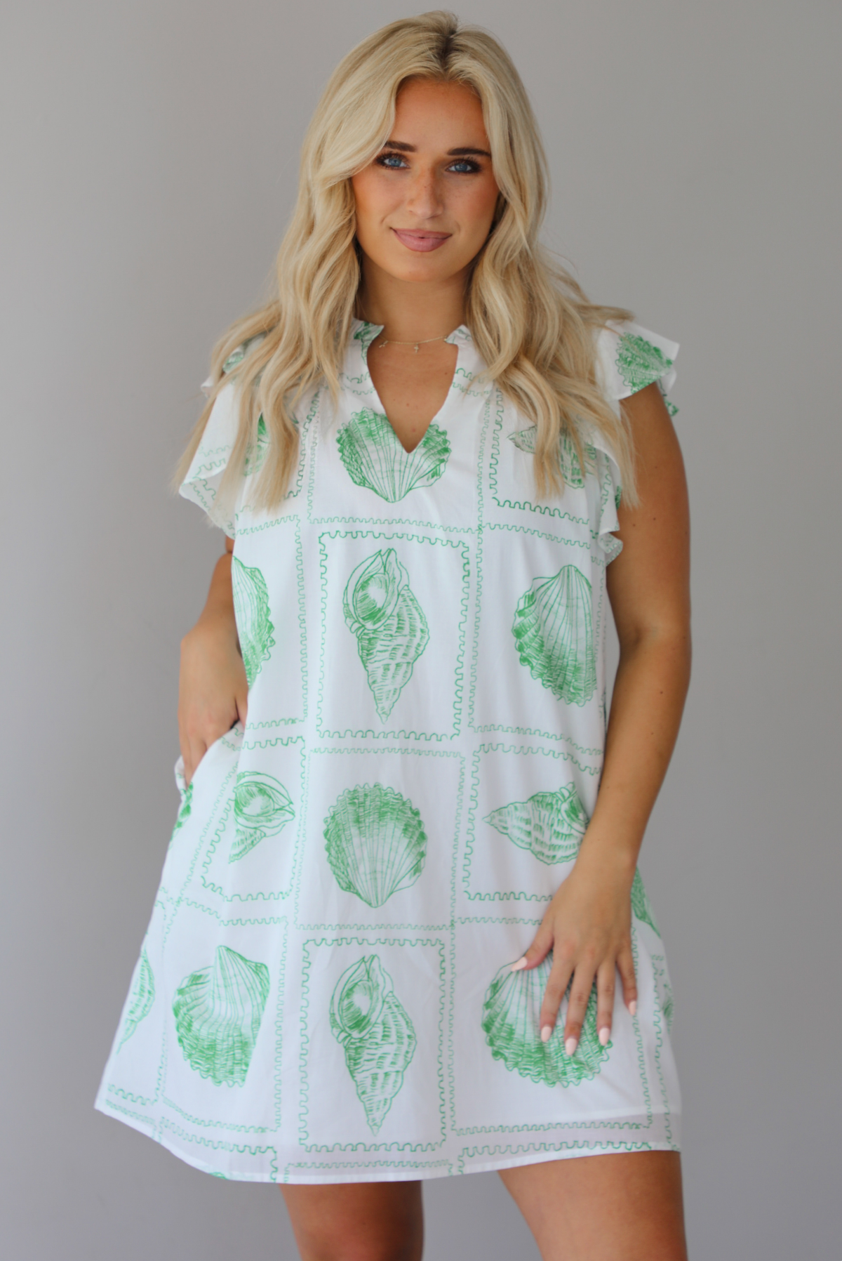 Woman wearing a white and green shell patterned dress with ruffled sleeves, functional pockets on either side, with a flowy skirt indoors against a white wall.