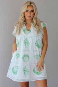 Woman wearing a white and green shell patterned dress with ruffled sleeves, functional pockets on either side, with a flowy skirt indoors against a white wall.