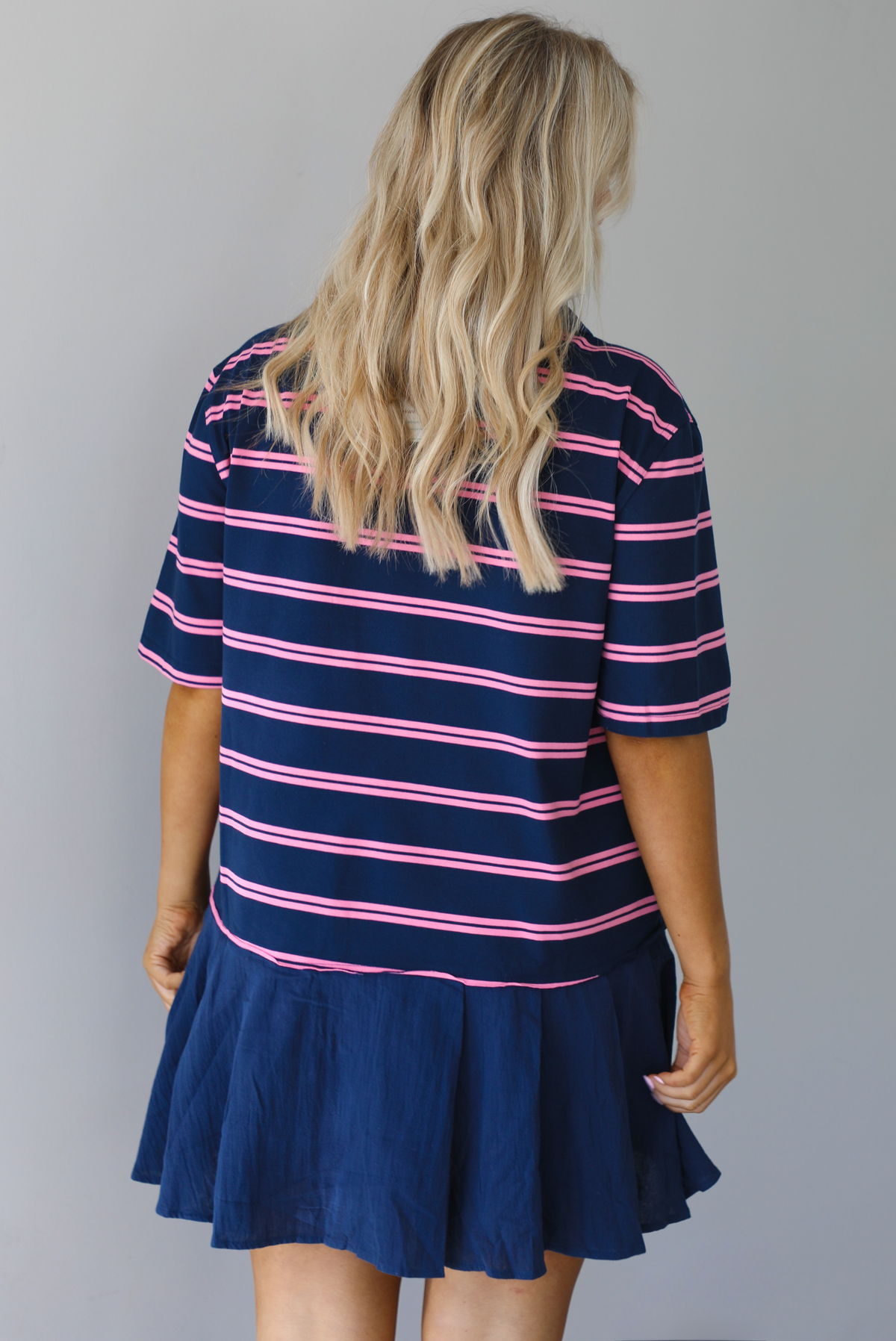 Woman wearing a navy dress with a pink striped design throughout, collared neckline, short sleeves, and a flowy bottom hem that's a different texture than the rest of the dress and is solid navy. The photo is taken indoors against a white wall and shows the back of the dress.