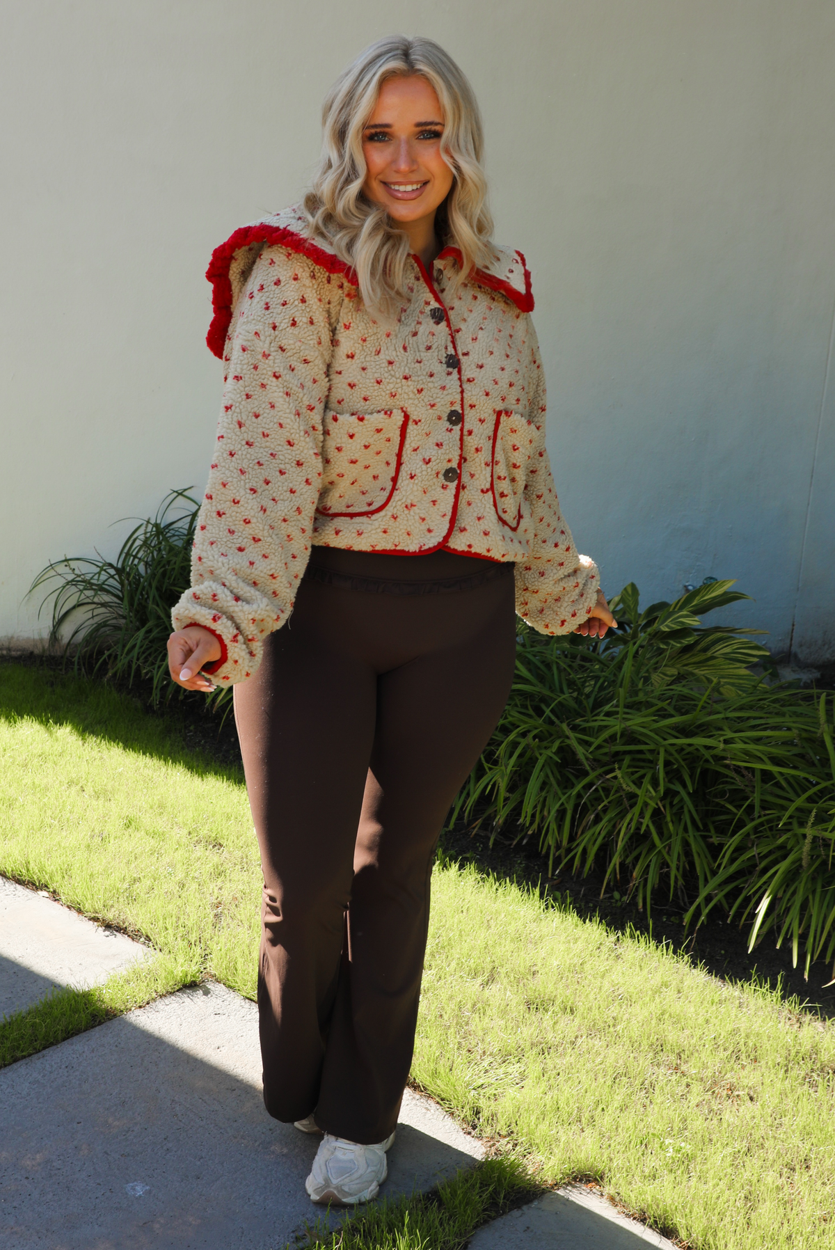 Woman model wearing a tan and red sherpa cardigan with a collared neckline and red trim with dark brown ahtleisure pants. The photo is taken outdoors with a white wall and greenery in the back. 