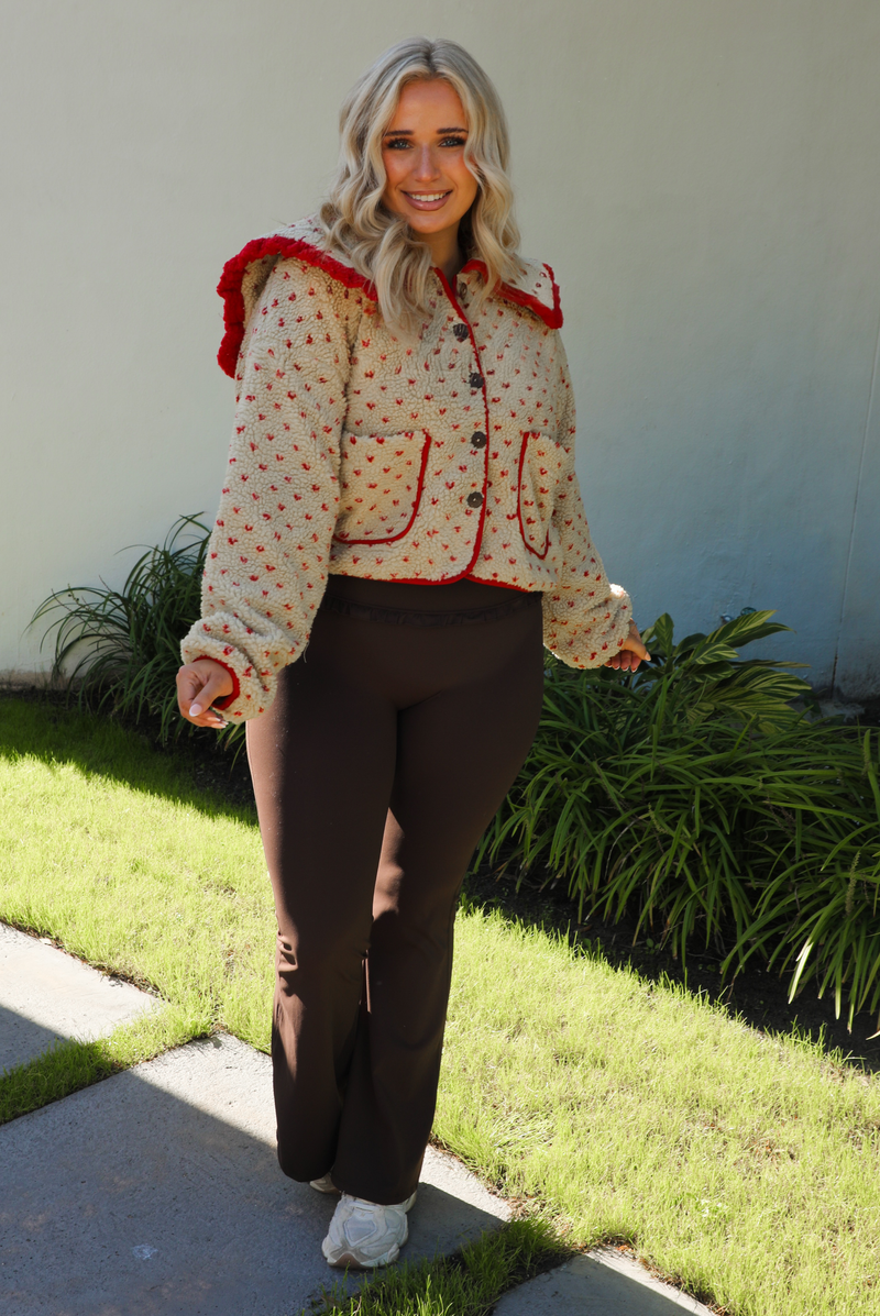 Woman model wearing a tan and red sherpa cardigan with a collared neckline and red trim with dark brown ahtleisure pants. The photo is taken outdoors with a white wall and greenery in the back. 