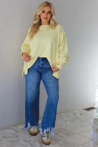 Woman wearing a pale yellow oversized sweater with dark wash denim and sneakers. The photo is taken indoors against a white wall with white marble flooring. 