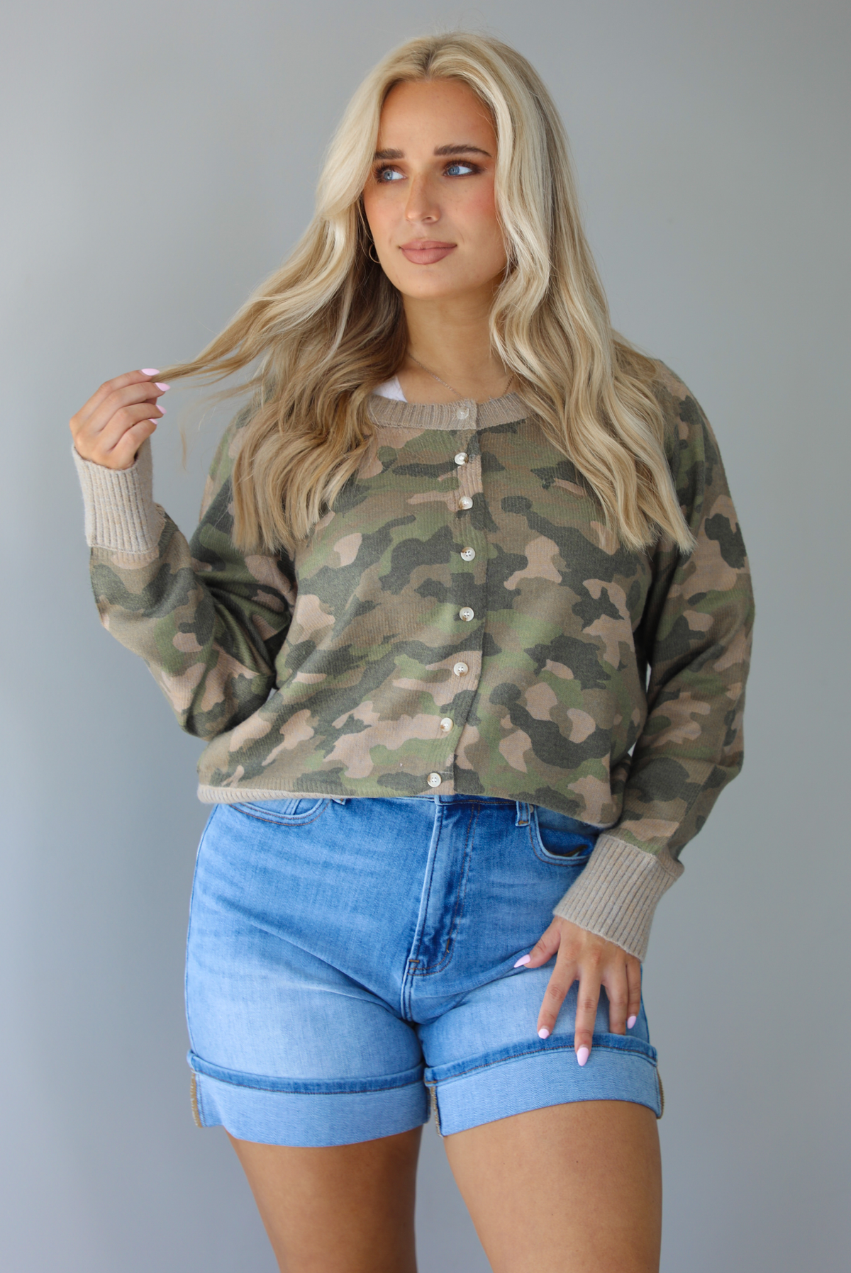 Woman wearing a camo cardigan with functional button closures in the front tucked into high waisted denim shorts. The photo is taken in front of a white wall.