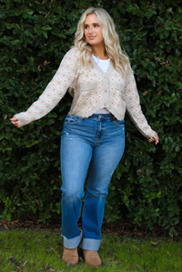 Woman model wearing a grey cardigan that has an orange and white floral print pattern throughout, long sleeves, an asymmetrical hem, and functional button closures in the front, with a long sleeved white top, medium wash denim jeans, and brown shoes in front of a green hedge outdoors. 