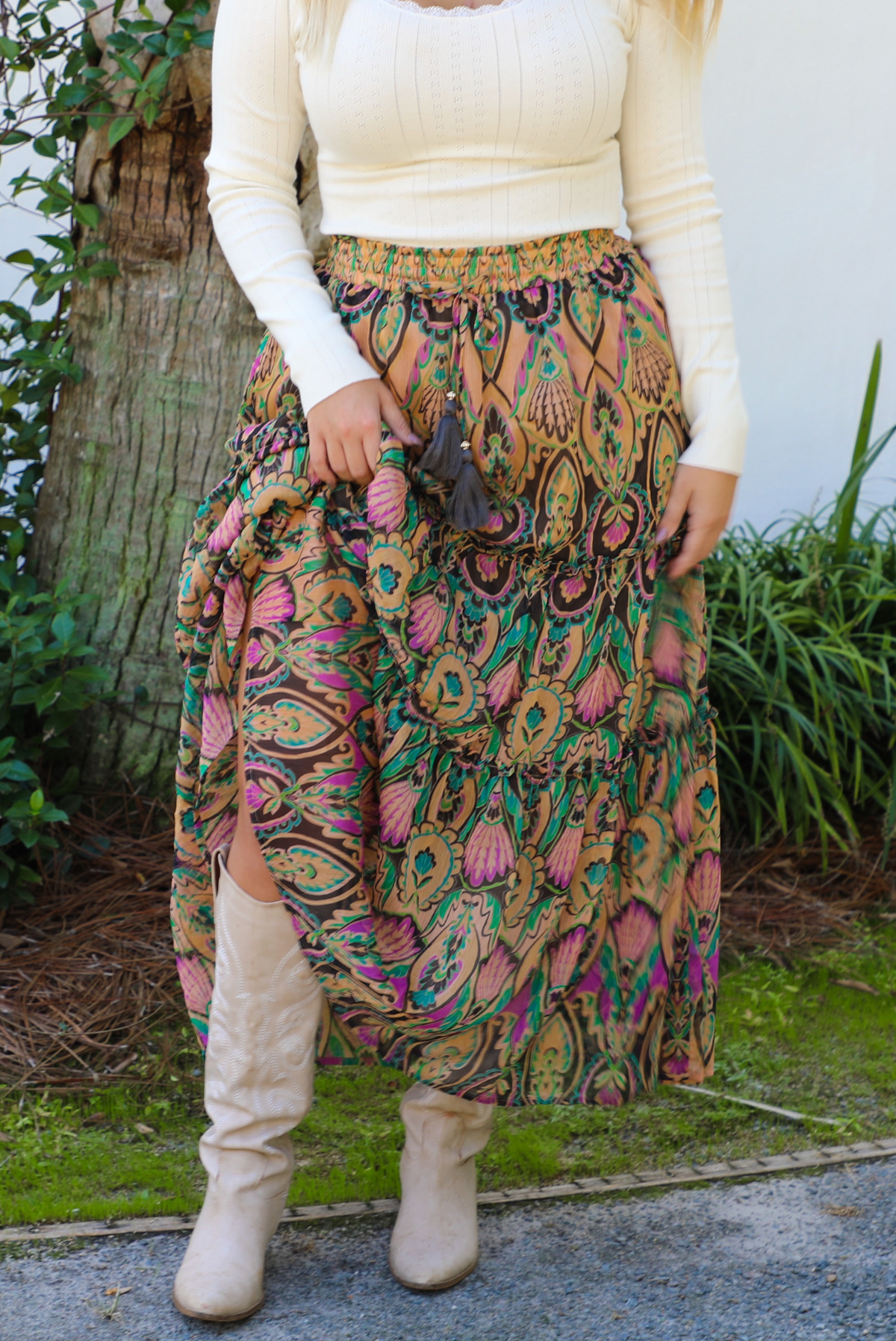 A closeup of a woman, whose face is not showing, wearing a long sleeved cream top and multi-colored mai length skirt paired with knee high sand colored ankle boots, standing outdoors with a white wall, tree, and other greenery in the background. 