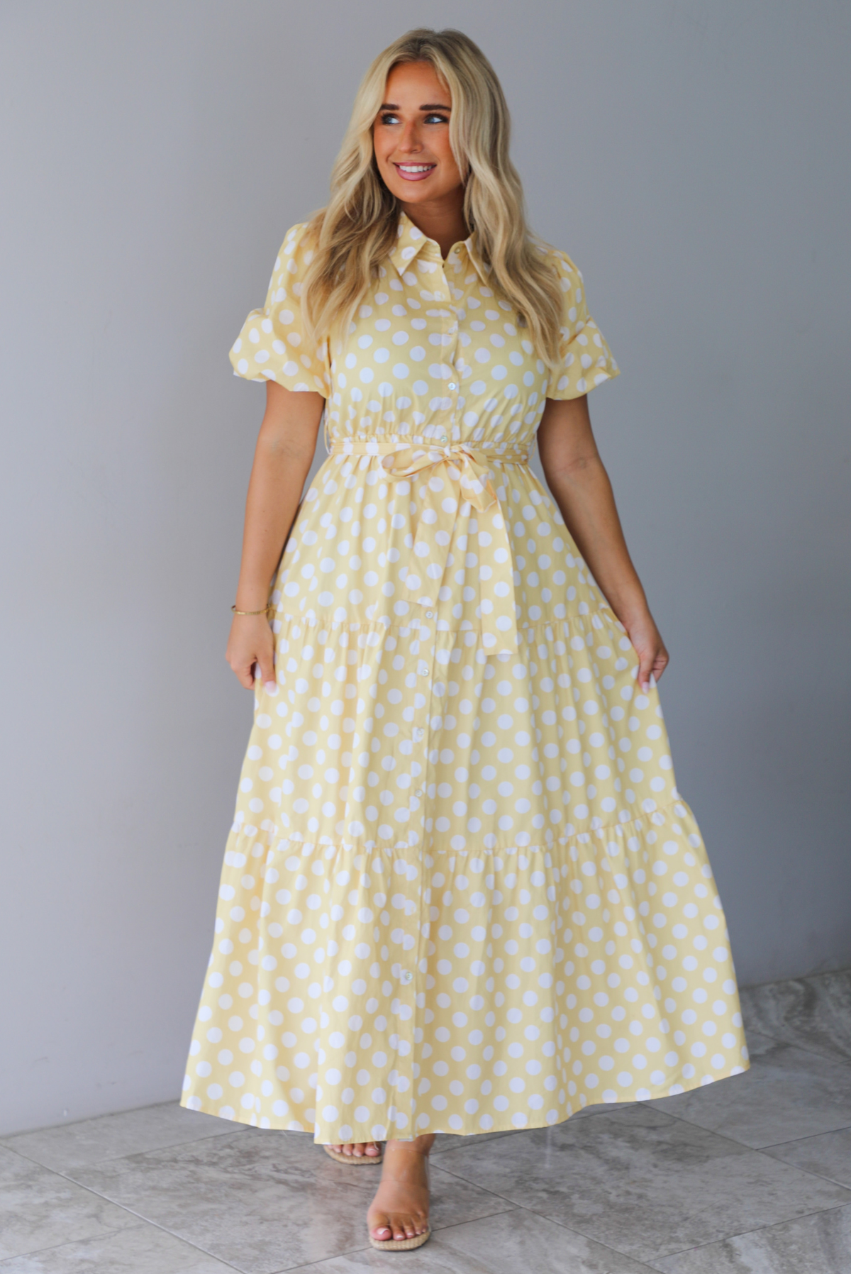 A woman wearing a yellow and white polka dot midi dress with puff sleeves and a tie waist and functional button closures along the front, standing indoors against a white wall with white marble flooring. 