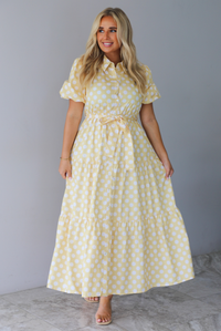 A woman wearing a yellow and white polka dot midi dress with puff sleeves and a tie waist and functional button closures along the front, standing indoors against a white wall with white marble flooring. 
