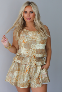 A woman wearing a halter neckline yellow and multi patchwork patterned romper that has a cinched waistline, a ruffled detail at the waistline, and functional pockets on either side, standing indoors against a white wall. 