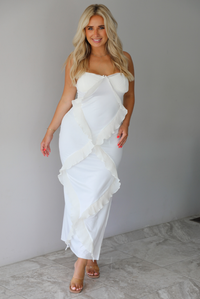 Woman wearing a white maxi length dress with ruffled details along the front indoors against a white wall with white marble flooring. 