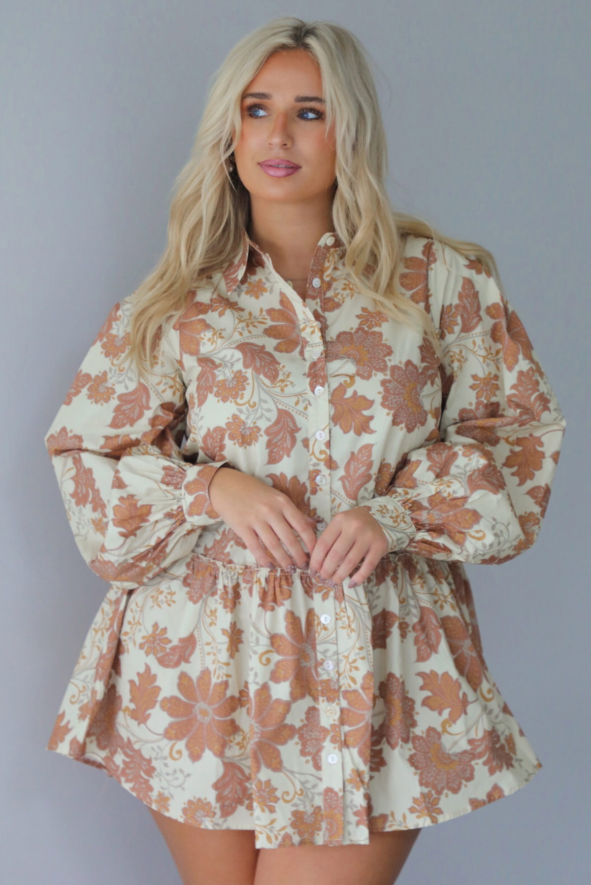 A woman wearing a cream dress with a tan fall floral print pattern throughout, collared neckline, functional button closures along the front, long sleeves that cinch tighter at the wrist, and a slightly flowy skirt, standing indoors against a white wall.