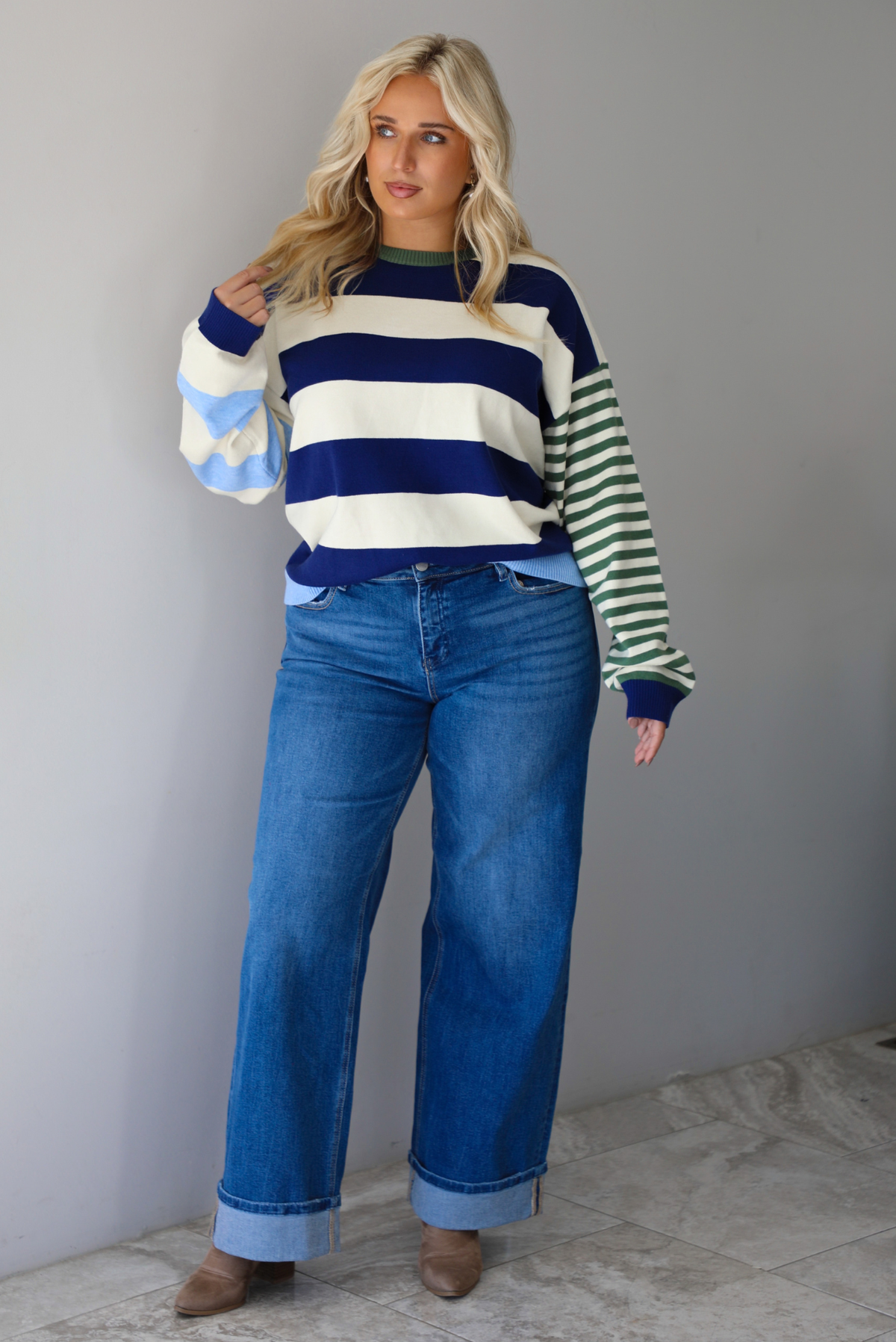 Woman wearing a navy, cream, blue, and green alternating striped design throughout, paired with dark denim jeans, standing indoors against a white wall with white marble flooring. 