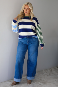 Woman wearing a navy, cream, blue, and green alternating striped design throughout, paired with dark denim jeans, standing indoors against a white wall with white marble flooring. 