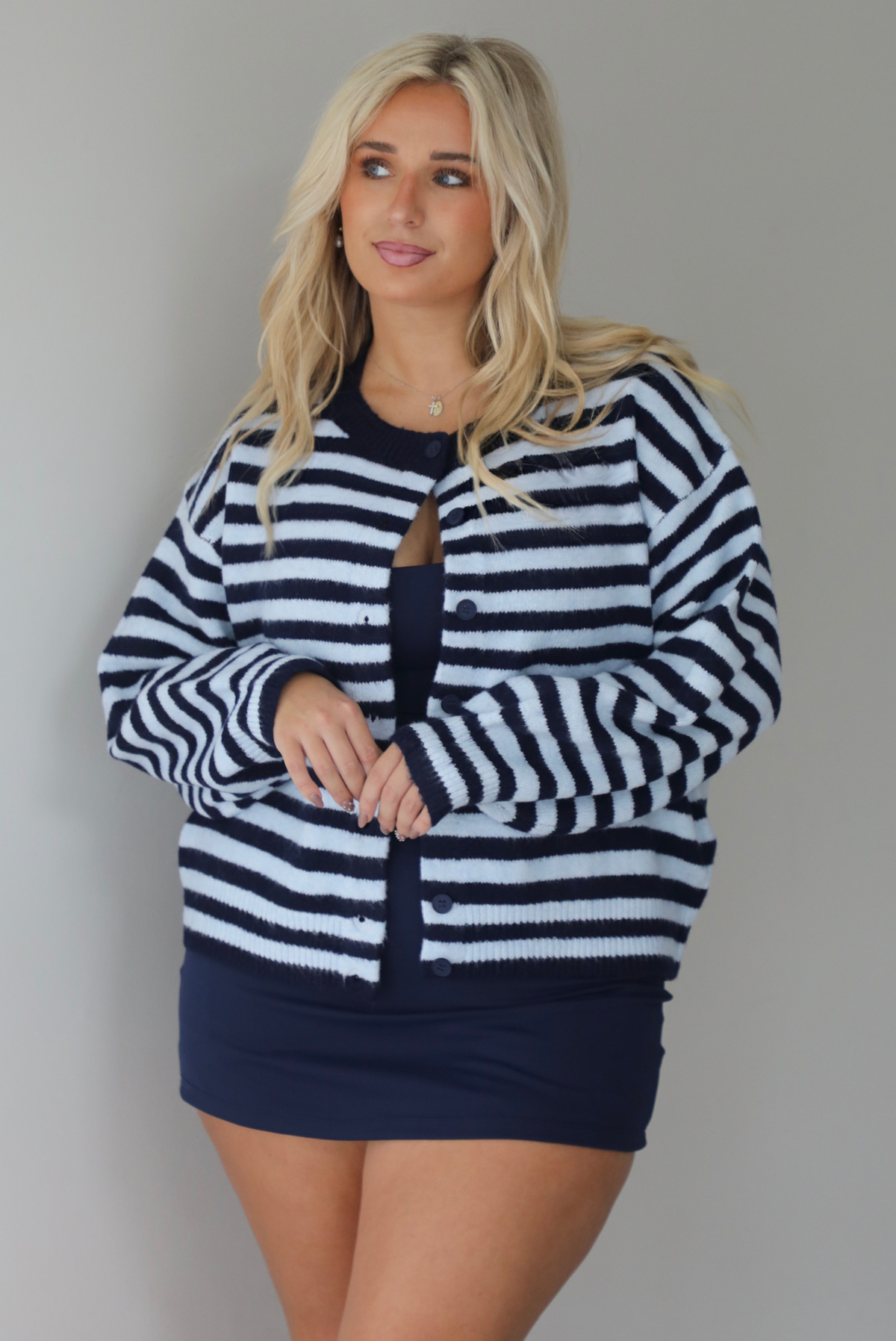 A woman wearing a navy and blue striped cardigan paired with a navy two piece set underneath standing indoors against a white wall.