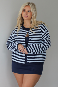 A woman wearing a navy and blue striped cardigan paired with a navy two piece set underneath standing indoors against a white wall.