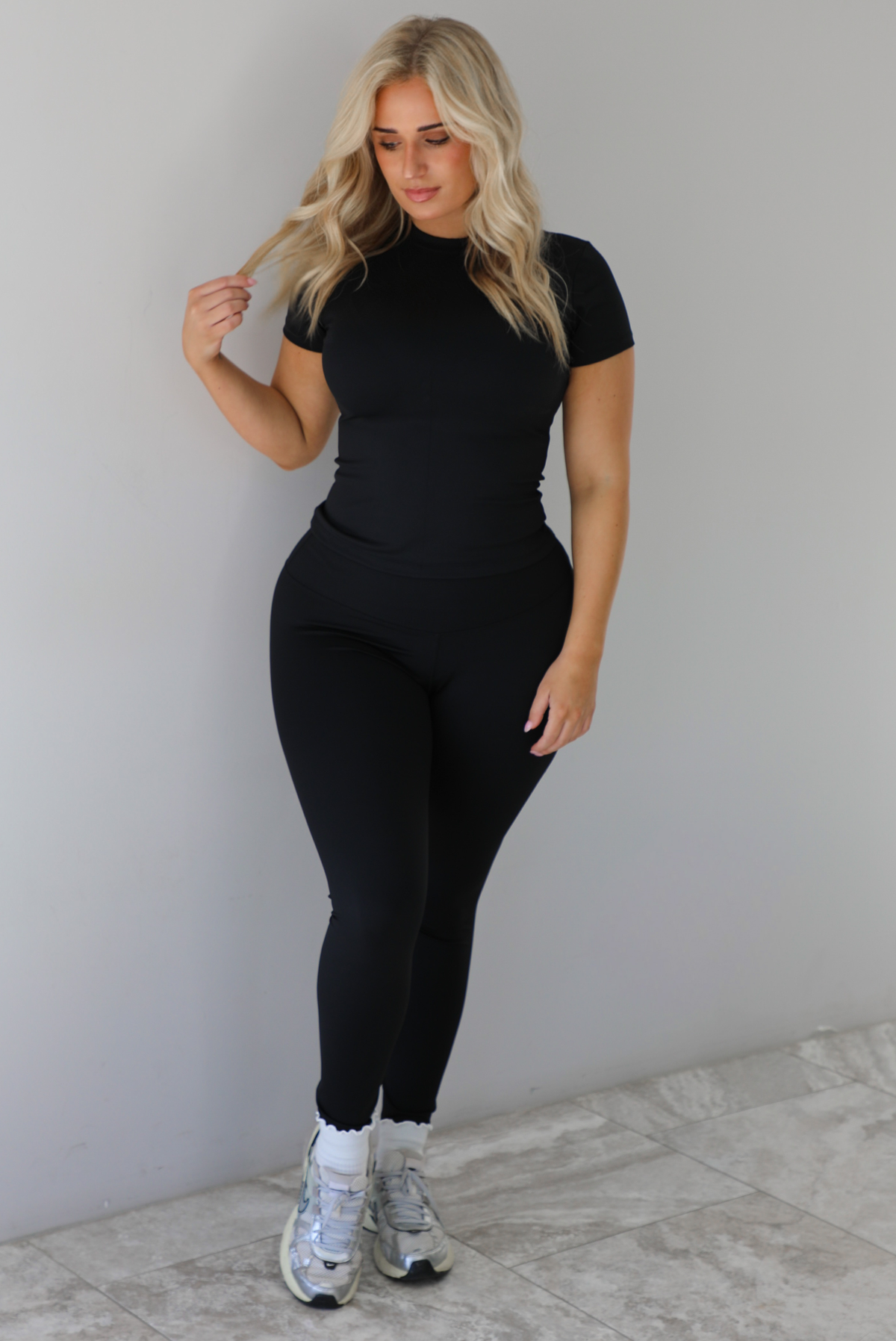 Woman wearing a fitted two piece black set featuring a short sleeved top and fitted leggings. The set is styled with a pair of white ankle socks and silver sneakers. The photo is taken indoors against a white wall with white marble flooring. 
