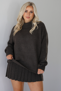 Woman wearing a dark brown sweater set featuring a turtle-neck sweater and a pleated skirt. The photo is taken front of a white wall.