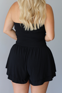 Woman wearing a black strapless romper with black boots indoors against a white wall. The photo shows a closeup of the back. 