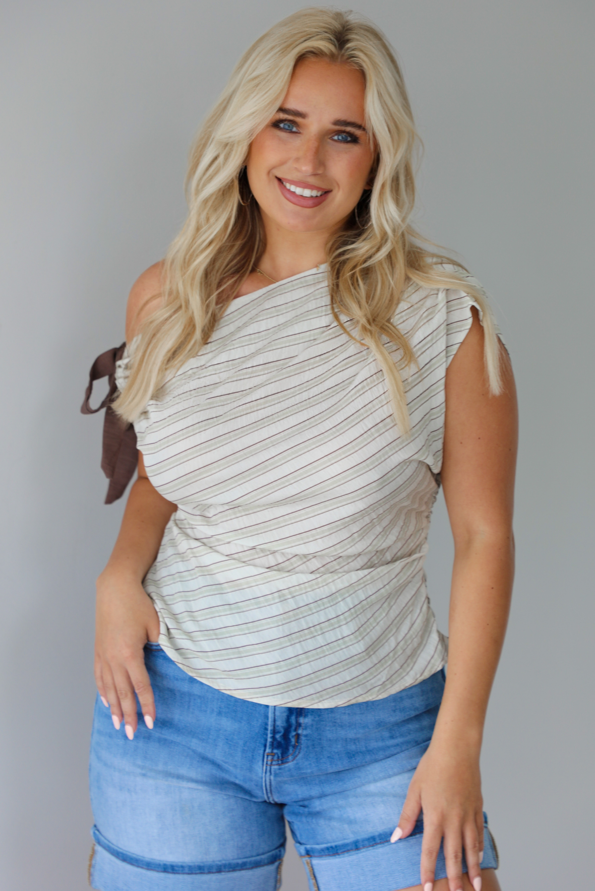Woman wearing a multi-colored striped top that has an off the shoulder design with a tie on the right side with medium wash denim shorts indoors against a white wall.