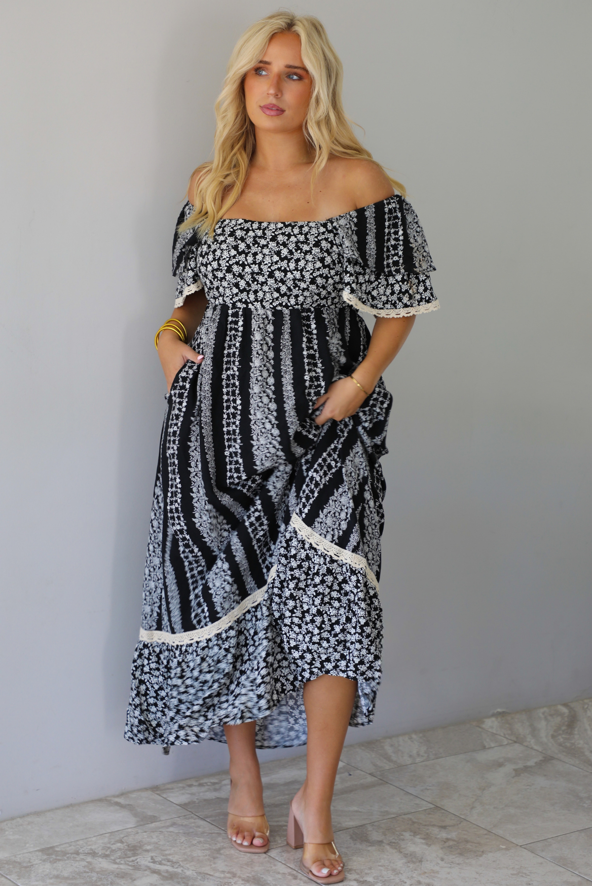 A woman wearing a black and white maxi dress with a white floral print that alternates between a cluster appearance and a striped appearance, with off the shoulder sleeves and a flowy skirt, standing indoors against a white wall with white marble flooring. 