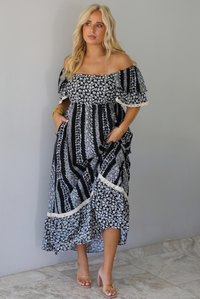 A woman wearing a black and white maxi dress with a white floral print that alternates between a cluster appearance and a striped appearance, with off the shoulder sleeves and a flowy skirt, standing indoors against a white wall with white marble flooring. 