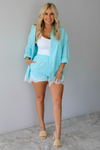 A woman stands indoors against a white wall on white marble flooring wearing a light aqua blue matching lounge set consisting of a long open-front button-down and shorts with white lace trim, over a white tank top and beige slide heeled sandals.