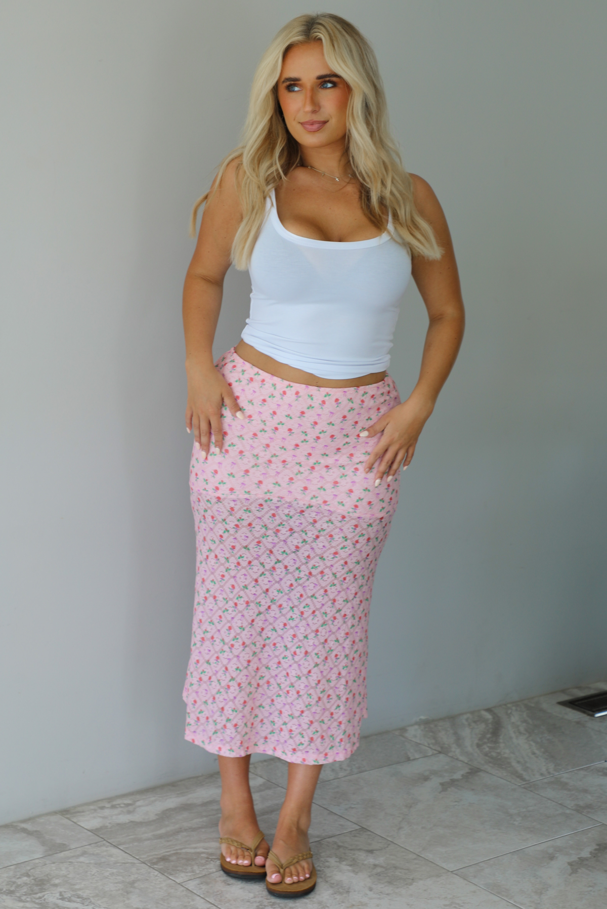 Woman wearing a white tank and pink midi length skirt that has a repeating floral pattern throughout indoors against a white wall with white marble flooring. 