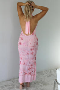 Woman wearing a formal pink maxi dress that has a plunging slouchy neckline and floral rhinestone details throughout. The photo is taken indoors against a white wall with white marble flooring and shows the back of the dress which has an open back design and halter tie neckline. 