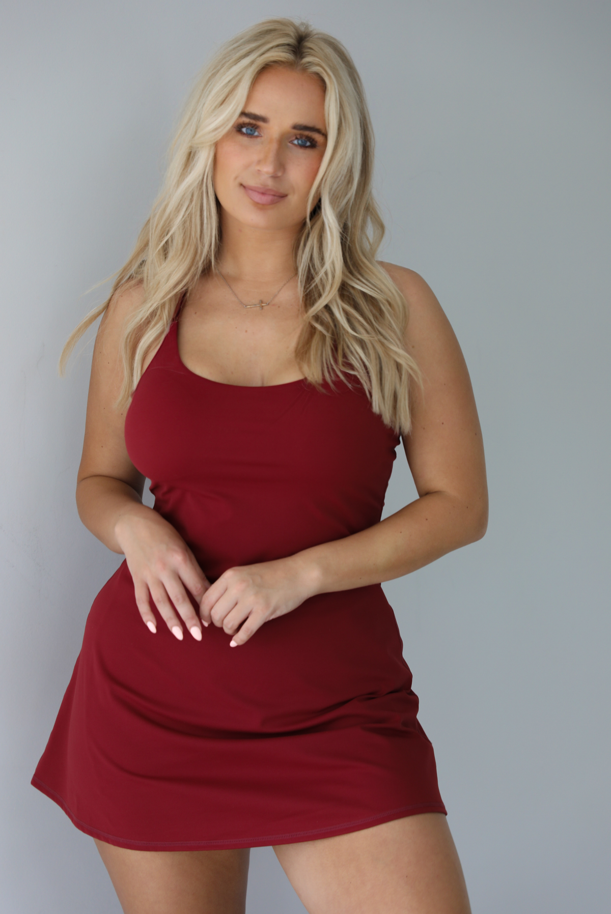 Woman wearing a maroon athleisure dress with thin straps and a relaxed fit indoors against a white wall.