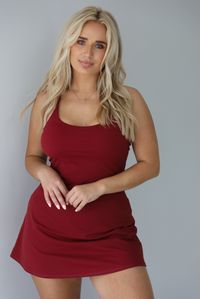 Woman wearing a maroon athleisure dress with thin straps and a relaxed fit indoors against a white wall.