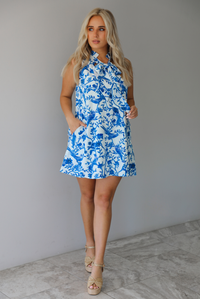 Woman wearing a white and blue print dress with a tree branch and bird pattern throughout indoors against a white wall with white marble flooring. 