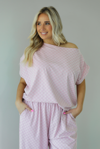 Woman wearing a pink checkered set featuring a slouchy top and pants. The photo is taken indoors against a white wall and shows a closeup of the top, which is worn off the shoulders and has a relaxed fit.