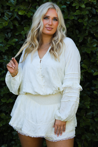Woman model wearing an off-white romper outdoors in front of greenery. 
