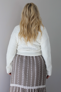 A woman shown from the back wearing an ivory v-neck sweater paired with a taupe and white embroidered maxi skirt, standing indoors against a white wall.