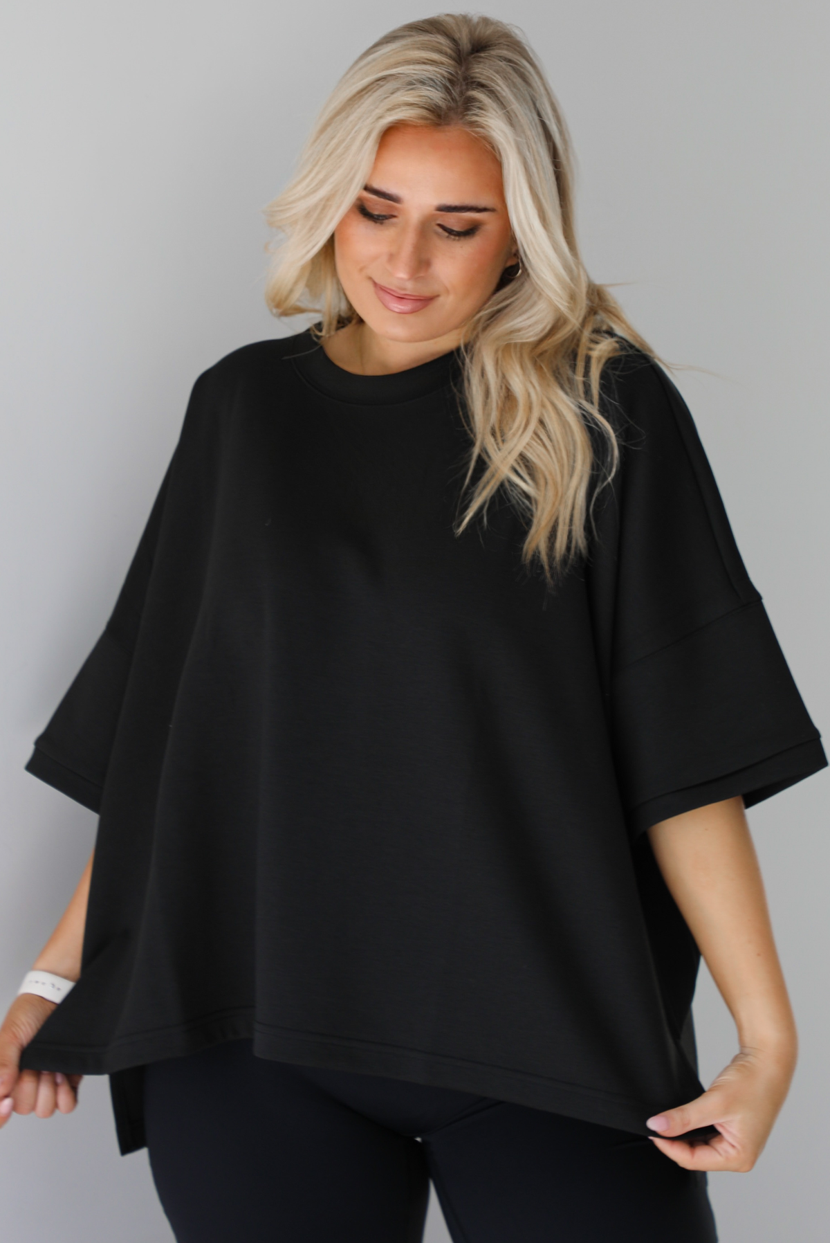 Woman wearing a black oversized box tee with longer short sleeves, a high-low design, and split hem details on either side. The tee is paired with black flared leggings. The photo is taken indoors against a white wall.