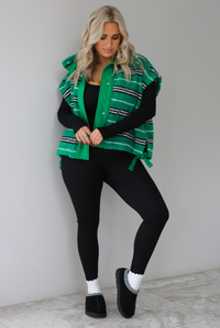 Woman wearing a green sherpa vest with black and white stripes that has an oversized fit, with a long sleeved, fitted, full body jumpsuit, with white slouchy socks and black ugg boot slides. The photo is taken indoors against a white wall with white marbled flooring. 