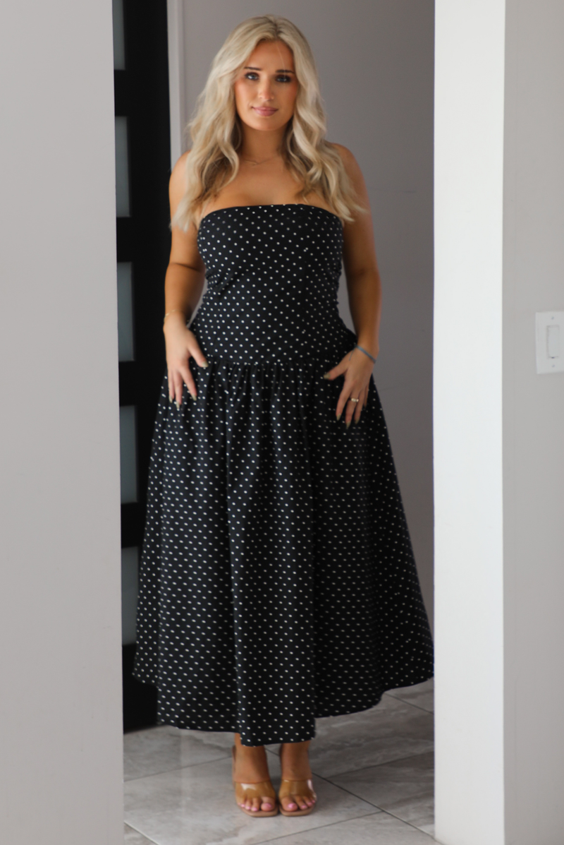 Woman model wearing a strapless/sleeveless, black with white polka dots, maxi length dress indoors. It has a fitted bodice with a flowy skirt. 