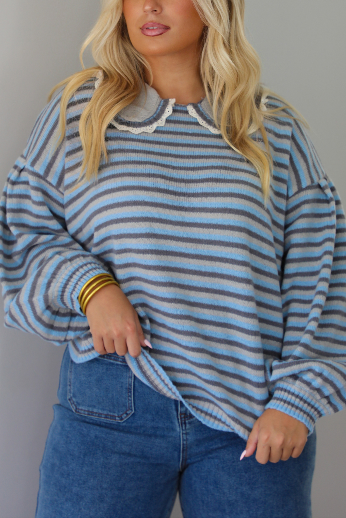 A closeup of a woman modeling a light blue and navy striped long-sleeve sweater with a white ruffled collar, paired with wide-leg blue jeans and brown boots, standing indoors against a white wall.