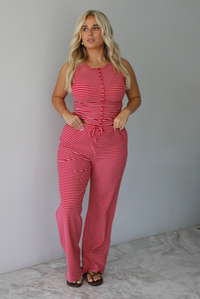 A woman modeling a red and white striped sleeveless set consisting of a tank top with non-functional button closures in the front and high waisted pants with a drawstring waistline and pockets on either side indoors against a white wall with white marble flooring. 