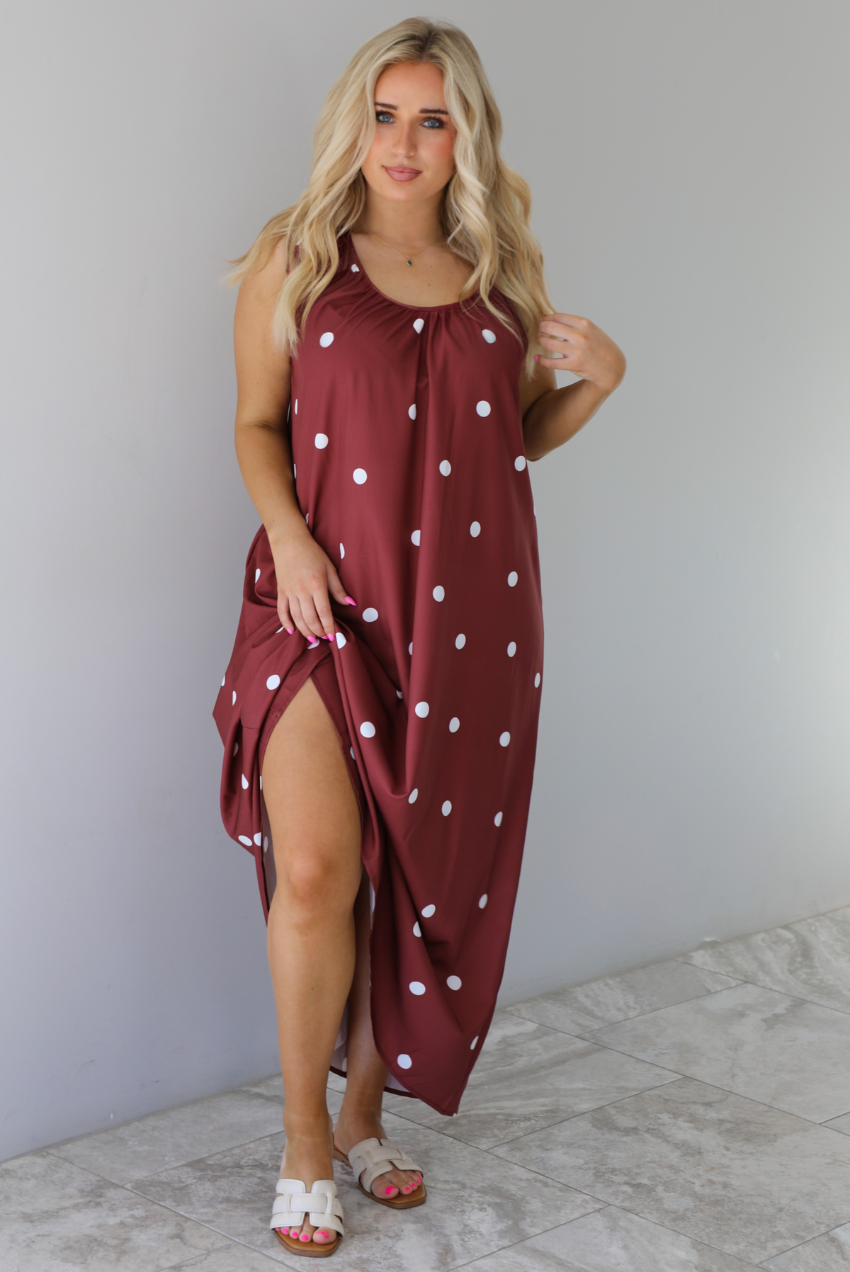 Woman wearing a red brown maxi length dress that has a white polka dot print pattern throughout. The dress is paired with sand sandals. The photo is taken indoors against a white wall with white marble flooring. 
