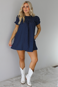 Woman wearing a navy baby-doll cut dress with a white polkadot print pattern throughout. The dress has puffed short sleeves and functional pockets on either side. The dress is paired with white cowboy boots. The photo is taken indoors against a white wall with white marble flooring. 
