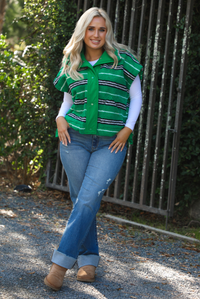 Woman model wearing a green, black, and white striped vest that has a sherpa texture to it, functional button closures in the front, and tie closures on either side, It is paired with a white long sleeved top, a medium wash pair of denim jeans, and tan ugg boots. The photo is taken outdoors with a metal gate and greenery in the back.
