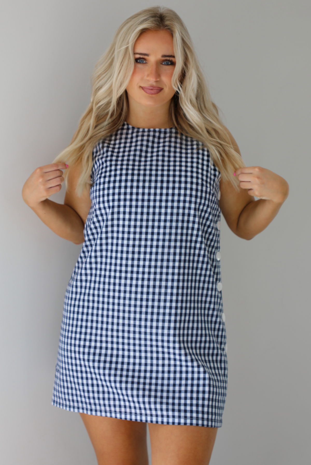 Woman wearing a navy and white gingham print patterned dress that has non-functional button closures on the left side. The photo is taken indoors against a white wall.