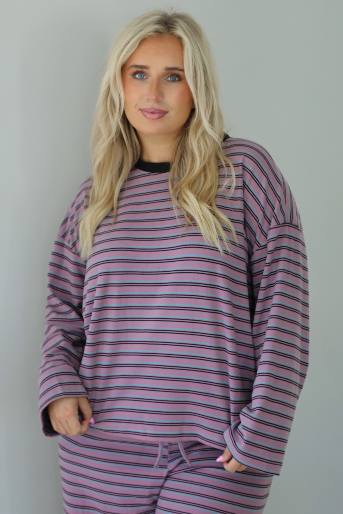 A woman wearing a purple matching set with a black and blue striped pattern featuring an oversized long sleeved top and high waisted flared hem pants, standing indoors against a white wall. The photo shows a closeup of the top, which has a thick black neckline detail. 