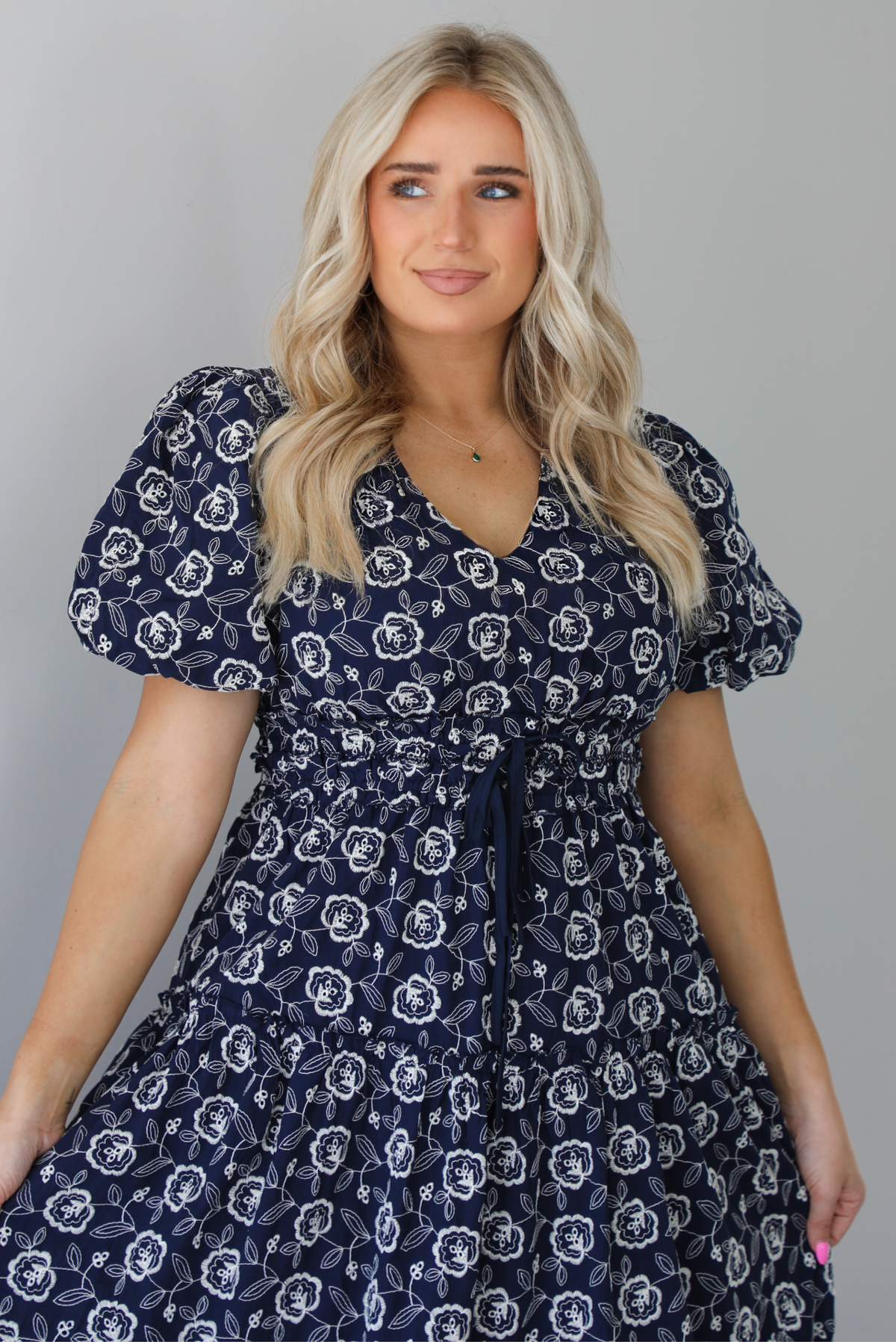 Woman wearing a navy dress that has a white embroidered floral print pattern throughout, a cinched waistline, and flowy maxi length skirt. The photo shows a closeup of the bodice. The photo is taken indoors against a white wall.