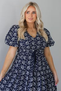 Woman wearing a navy dress that has a white embroidered floral print pattern throughout, a cinched waistline, and flowy maxi length skirt. The photo shows a closeup of the bodice. The photo is taken indoors against a white wall.