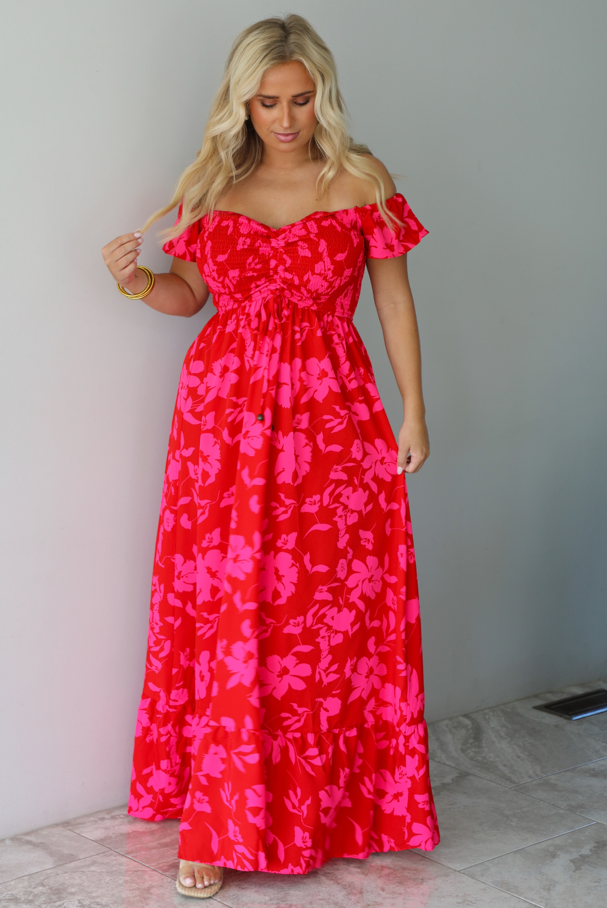 A woman modeling a bright red off-the-shoulder maxi dress with a vibrant pink floral pattern, standing indoors agains a white wall with white marble flooring. 