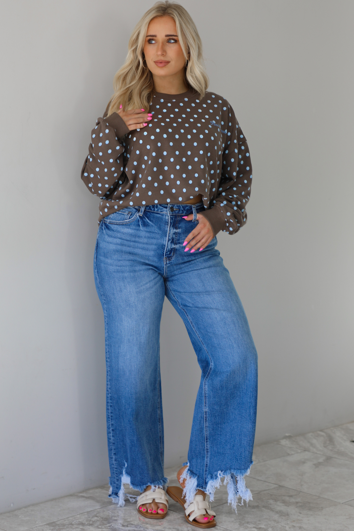 Woman wearing a brown sweater that has a blue polka dot print pattern throughout with blue embroidered writing on the left side that reads 'Trust The Process.' The sweater is paired with a medium wash denim. The photo is taken indoors against a white wall with white marble flooring.