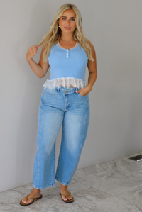 A woman wearing a light blue sleeveless top with a white lace hem and light blue high-waisted cropped jeans, standing indoors against a white wall with white marble flooring. 